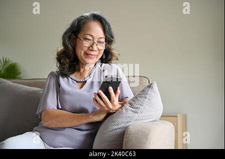 Bella e splendida donna asiatica anziana indossando occhiali con il suo smartphone per leggere una notizia online mentre si rilassa sul suo comodo divano. Foto Stock