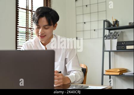 Bel giovane uomo d'affari asiatico o lavoratore maschile che lavora alla sua scrivania, gestendo le sue attività aziendali su un computer portatile. Foto Stock