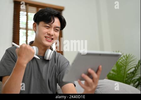 Un giovane asiatico bello e felice che utilizza il suo tablet digitale in salotto, riceve un'e-mail inaspettata, supera l'esame o riceve un'offerta di lavoro Foto Stock