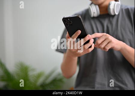 Giovane asiatico in abiti casual in salotto, utilizzando lo smartphone per inviare messaggi ai suoi amici, testando nuove applicazioni, ordinando cibo di consegna sul Foto Stock