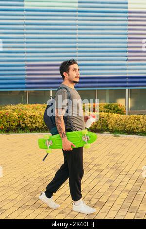 Giovane uomo che cammina per la città sul suo skateboard mentre beve caffè. Foto Stock