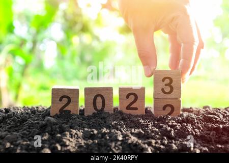 Mano che cambia anno 2022 a 2023 in blocchi di legno cubetti con pianta crescente all'alba. Anno nuovo 2023, Hope, Hello e Arrivederci Concept. Foto Stock