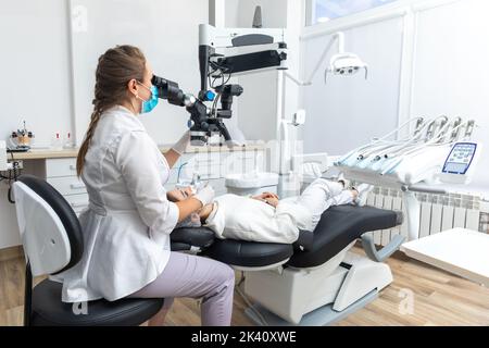 Dentista femmina che usa un microscopio dentale che tratta i denti del paziente presso la clinica dentale Foto Stock