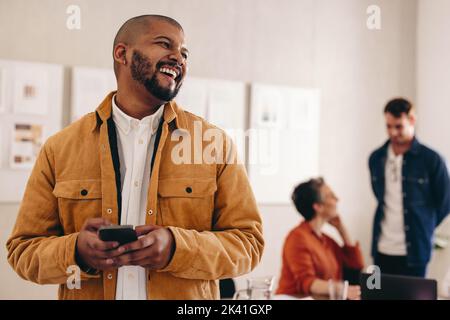 Buon uomo d'affari con un anello con setto che guarda lontano mentre tiene uno smartphone. Giovane uomo d'affari allegro che lavora in un ambiente di lavoro creativo e moderno wi Foto Stock