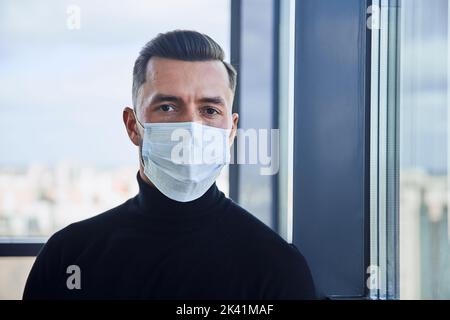 giovane uomo in una maschera protettiva in piedi vicino alla finestra. Foto Stock