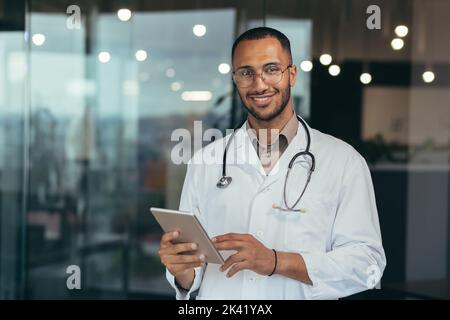 Ritratto di felice e riuscito dottore afroamericano che lavora all'interno di una clinica d'ufficio tenendo un computer tablet guardando la macchina fotografica e sorridendo indossando un cappotto bianco con stetoscopio. Foto Stock