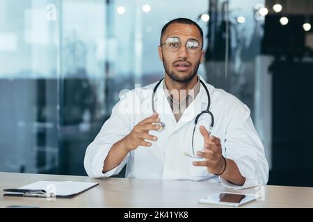 Medico serio e focalizzato in cappotto medico e stetoscopio parlando di videochiamata guardando la telecamera e consulenza paziente, uomo che lavora all'interno di clinica ufficio. Foto Stock