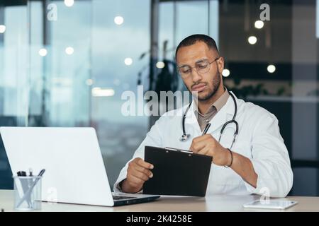 Medico ispanico serio e focalizzato in cappotto medico e stetoscopio dietro la carta, uomo in occhiali che lavora in clinica ufficio utilizzando il computer portatile. Foto Stock