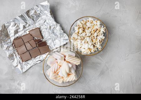 Popcorn, marshmallow pastello colorato, pezzi di barretta di cioccolato al latte rotto in ciotole di vetro su sfondo grigio. Ingredienti per cucinare dessert e dolci malsani. Vista dall'alto, spazio di copia Foto Stock