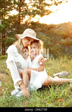 Donna morbida, allegra, sorridente in cappello e bambina bionda in abito bianco coccolante, abbracciante insieme sul prato Foto Stock