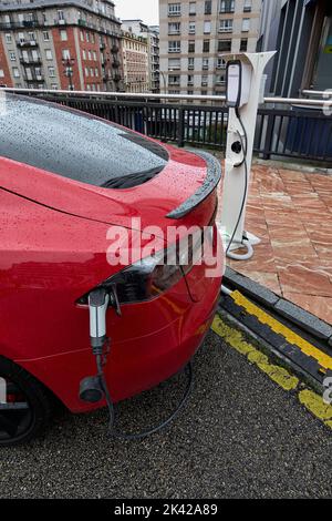 Auto elettrica rossa in una giornata di pioggia collegata con un adattatore a una stazione di servizio elettrica. Concetto di energia pulita come misura per rigenerare il pianeta Foto Stock