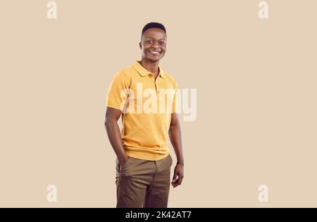Giovane uomo afroamericano sorridente che posa con una mano in pantaloni tascabili si erge su sfondo beige Foto Stock