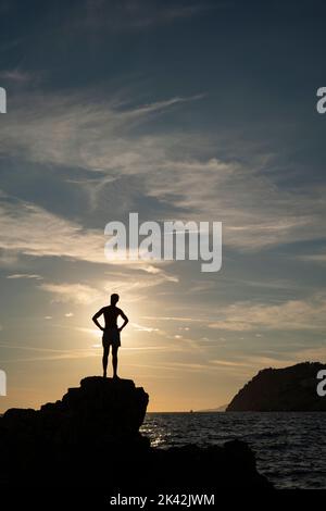 silhouette di un giovane uomo in piedi su una scogliera al tramonto Foto Stock