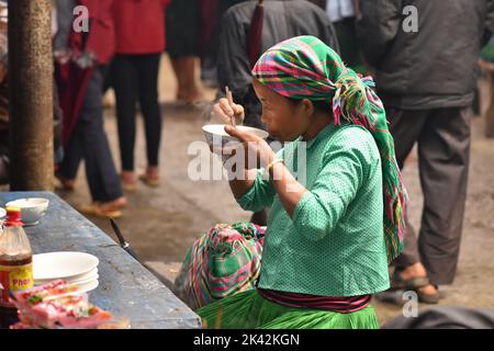 Donna di un gruppo etnico vietnamita che indossa abiti colorati tradizionali, pranzando nel mercato coperto della cittadina di Meo Vac, Vietnam. Foto Stock