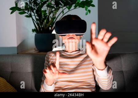 Giovane donna in maglione a righe e occhiali VR esplora un gioco interessante con espressione entusiasta Foto Stock