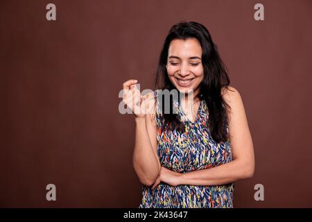 Donna indiana allegra ridendo con gli occhi chiusi ritratto, donna felice tenendo la mano piegata, schiaffo dita. Persona sorridente che posa la vista frontale studio medio girato su sfondo marrone Foto Stock