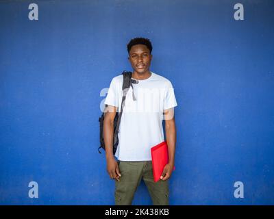 Ritratto di un giovane studente afro-americano che guarda la fotocamera con uno zaino e una cartella. Ritorno a scuola, scuola superiore, università Foto Stock