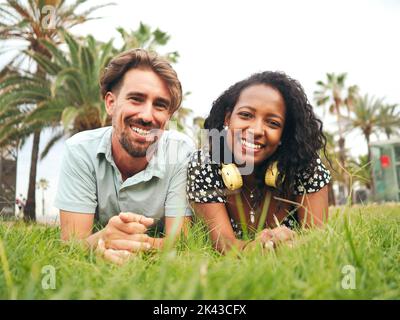 Giovane coppia eterosessuale multietnica sorridente sdraiata sull'erba in un parco Foto Stock