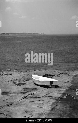 Una barca a remi bianca abbandonata sulla costa rocciosa del Faro di Nubble a Cape Neddick, Maine. L'immagine è stata catturata in una pellicola analogica in bianco e nero. Foto Stock