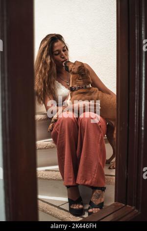 Bella giovane latina abbracciando il suo cane mostrando il suo affetto sulle scale della sua casa. Amore per gli animali. Foto Stock