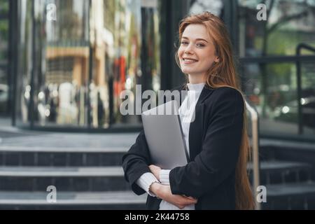 Sicuro modello di donna d'affari che tiene il laptop che guarda la macchina fotografica sorridente toothy in piedi fuori all'esterno vicino all'azienda. Donna caucasica, persona d'affari Foto Stock