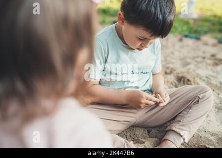 Concentrato adorabile ragazzo studiando una pietra che trovò nella sabbia del giardino. Stile di vita all'aperto. Giardino di casa. Sviluppo del bambino. Natura estiva. Foto Stock