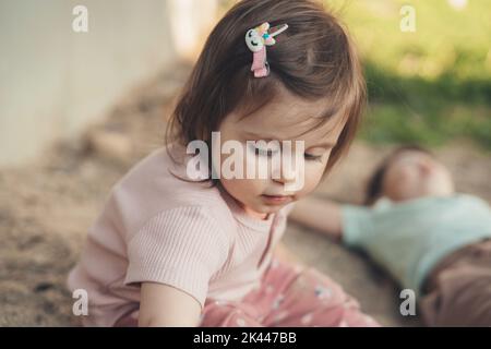 Primo piano ritratto di una bambina seduta nella sabbia in giardino con suo fratello, esplorando. Attività estive per bambini. Giorno estivo di sole Foto Stock