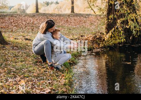 Madre con bambini pesca con un bastone al bordo di un lago nel parco autunnale. Natura selvaggia e divertimento in acqua durante le vacanze autunnali. Campeggio e pesca Foto Stock