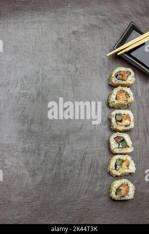 Sushi sono disposti verticalmente in una fila. Ciotola con salsa di soia e bastoncini. Spazio di copia. Giacitura piatta. Sfondo marrone. Foto Stock