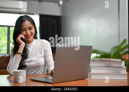 Bella e felice giovane ragazza asiatica freelancer nel suo ufficio a casa, al telefono con il suo cliente per fare un affare di affari. Foto Stock