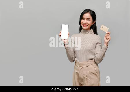 Bella bella giovane asiatica femmina con uno smartphone bianco schermo mockup e una carta di credito, in piedi isolato su sfondo grigio studio. Onlin Foto Stock