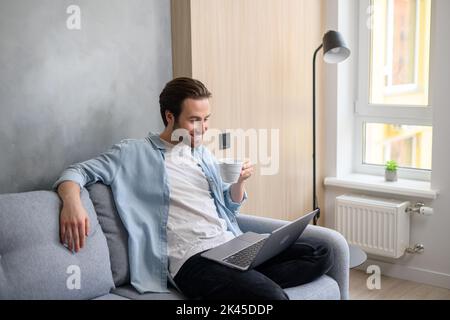 Sereno giovane 30s caucasico che utilizza il computer portatile in un'atmosfera rilassata casa, seduto sul divano e digitando, navigare sul Web, guardare film. Uomo spensierato bere bevande Foto Stock