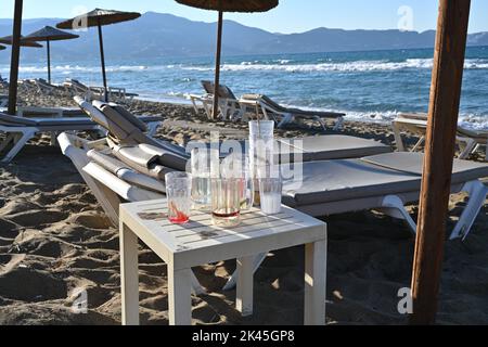 Tagliare la nostra di una spiaggia di sabbia di una località di vacanza con sedie a sdraio in plastica, bicchieri da cocktail vuoti e ombrelloni da spiaggia di paglia. La costa è all'orizzonte. Foto Stock