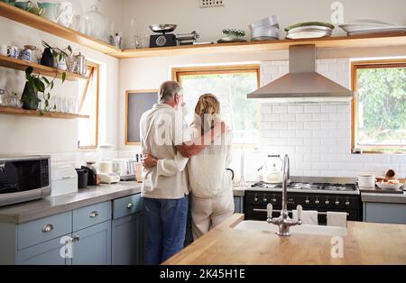 Abbraccio di coppia, caffè del mattino e anziani che hanno la colazione in cucina mentre si guarda la natura all'aperto. Uomo e donna anziani che abbraccia il tè Foto Stock