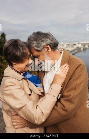 donna anziana che abbraccia con marito bearded in cappotto e sorridente vicino al fiume, immagine di scorta Foto Stock