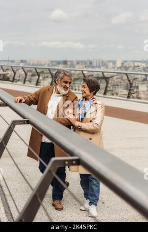 donna anziana felice abbracciando marito bearded vicino ponte guardia rail, immagine stock Foto Stock