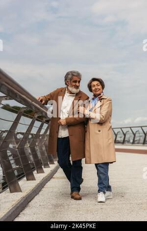 intera lunghezza di donna anziana felice che abbraccia il marito bearded e allegro vicino alla guida di guardia del ponte, immagine di scorta Foto Stock