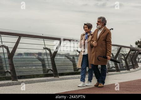 lunghezza intera di donna anziana sorridente che cammina con il marito bearded che tiene l'ombrello vicino alla guida di guardia del ponte, immagine di riserva Foto Stock