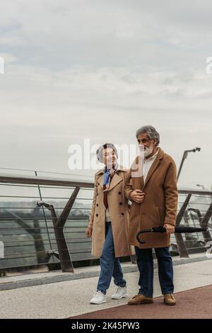 lunghezza intera di donna anziana allegra che cammina con il marito bearded che tiene l'ombrello vicino alla guida di guardia del ponte, immagine di riserva Foto Stock