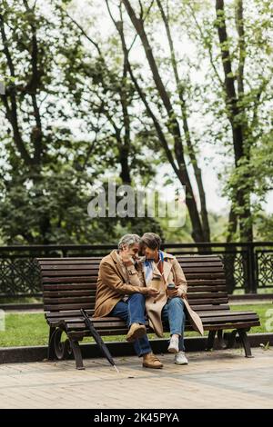 felice coppia in pensione in cappotti che tengono le tazze di carta e seduto sulla panchina nel parco, immagine di scorta Foto Stock