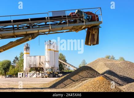 Un nastro trasportatore sopra mucchi di ghiaia e una pala gommata parcheggiata accanto a un silo di sabbia in una cava in una giornata di sole. Foto Stock