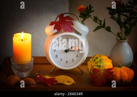 Caduta indietro, cambiamento di ora su una sveglia bianca dopo l'ora legale, candela, foglie e zucche come decorazione autunnale, fuoco selezionato Foto Stock