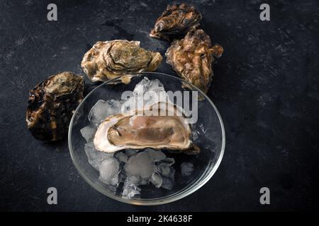 Ostriche aperte su ghiaccio tritato su un piatto di vetro e ostriche chiuse su uno sfondo rustico scuro, spazio copia, vista dall'alto dal fuoco selezionato Foto Stock