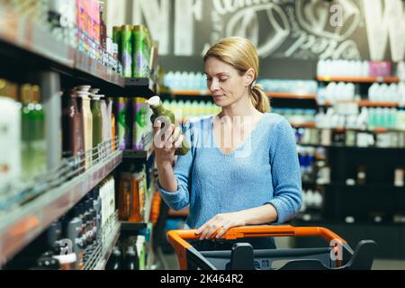 Una giovane donna sta in piedi in un deposito nel reparto dei prodotti chimici domestici con un carrello per le merci, sceglie lo shampoo per i capelli, tiene uno verde nella sua mano, legge la composizione del gel, shampoo. Foto Stock