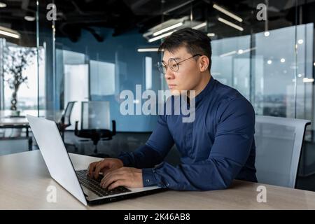 Uomo d'affari serio e concentrato che lavora con il laptop, uomo in occhiali e camicia all'interno dell'edificio ufficio, boss asiatico che scrive sul computer. Foto Stock