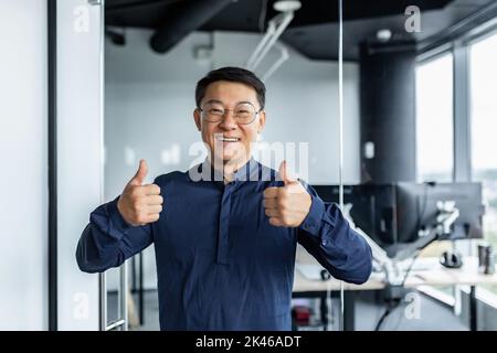 Ritratto di un uomo d'affari asiatico di successo, uomo che lavora in un ufficio moderno guardando la fotocamera e sorridendo, broker trader mostrando pollici in su. Foto Stock