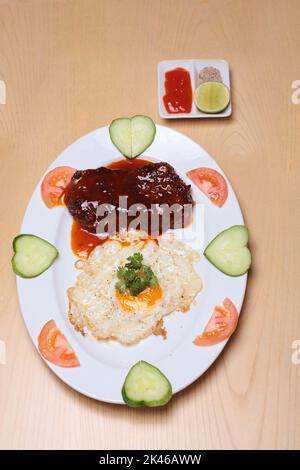 Bistecca di manzo con uova fritte e verdure sono poste su un piatto bianco, su un tavolo di legno, servito con salsa di peperoncino, pepe di sale e limone Foto Stock