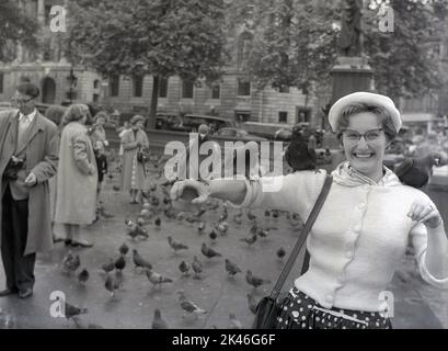 1958, storici piccioni seduti su un braccio femminile a Trafalgar Square, Westminster, Londra, Inghilterra, Regno Unito. In mano una scatola di piselli che ha portato da un chiosco che li ha venduti ai visitatori. Foto Stock