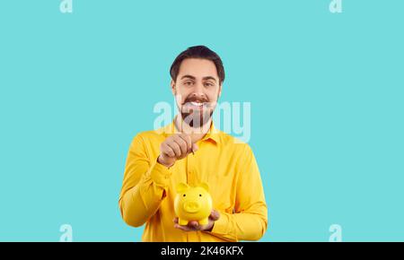 Giovane uomo caucasico allegro tiene la banca di piggy in mano a risparmiare soldi, si trova in studio turchese Foto Stock