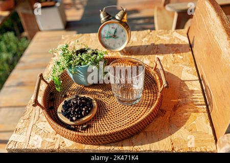 Una tazza di vetro vuota con chicchi di caffè, pianta in vaso su rattan vassoio di servizio su tavolo di legno nel pomeriggio luce solare Foto Stock